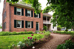 The Wylie House Museum, an old brick, two-story home, is shown surrounded by grass, bushes, flowers, and trees.